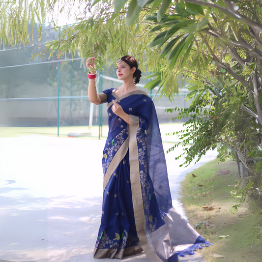 Navy Blue Zari Weaving Linen Silk Saree
A woman models a vibrant Navy Blue Linen Silk Saree with delicate gold/silver Zari woven motifs and a wide metallic silver border. She wears a beige blouse, red and white bangles, and a traditional headpiece, posing outdoors under a tree.