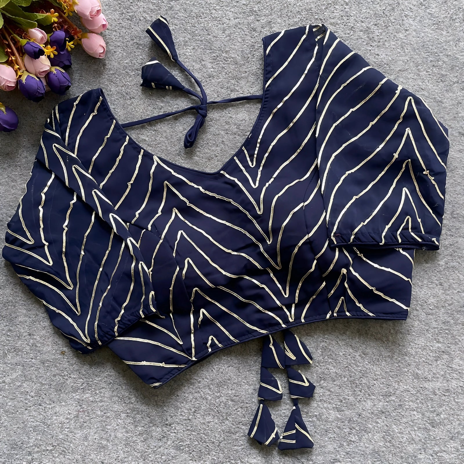 Navy blouse beautifully adorned with gold gota patti zigzag embroidery, three-quarter sleeves, and a stylish back tie-up with matching triangular tassels. Displayed flat on a grey textured surface beside pink and purple flowers, showcasing its festive and sophisticated ethnic design.