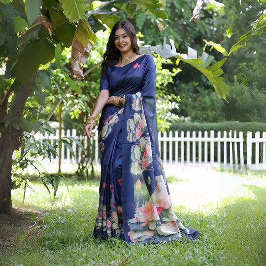 A woman models a stunning Deep Navy Blue Soft Tussar Silk Saree. The saree features a large-scale digital print of colorful floral and lotus motifs in pink, white, and green on the pallu and lower drape. She wears a matching blue blouse and heavy gold Kundan jewelry, posing outdoors in a garden setting.