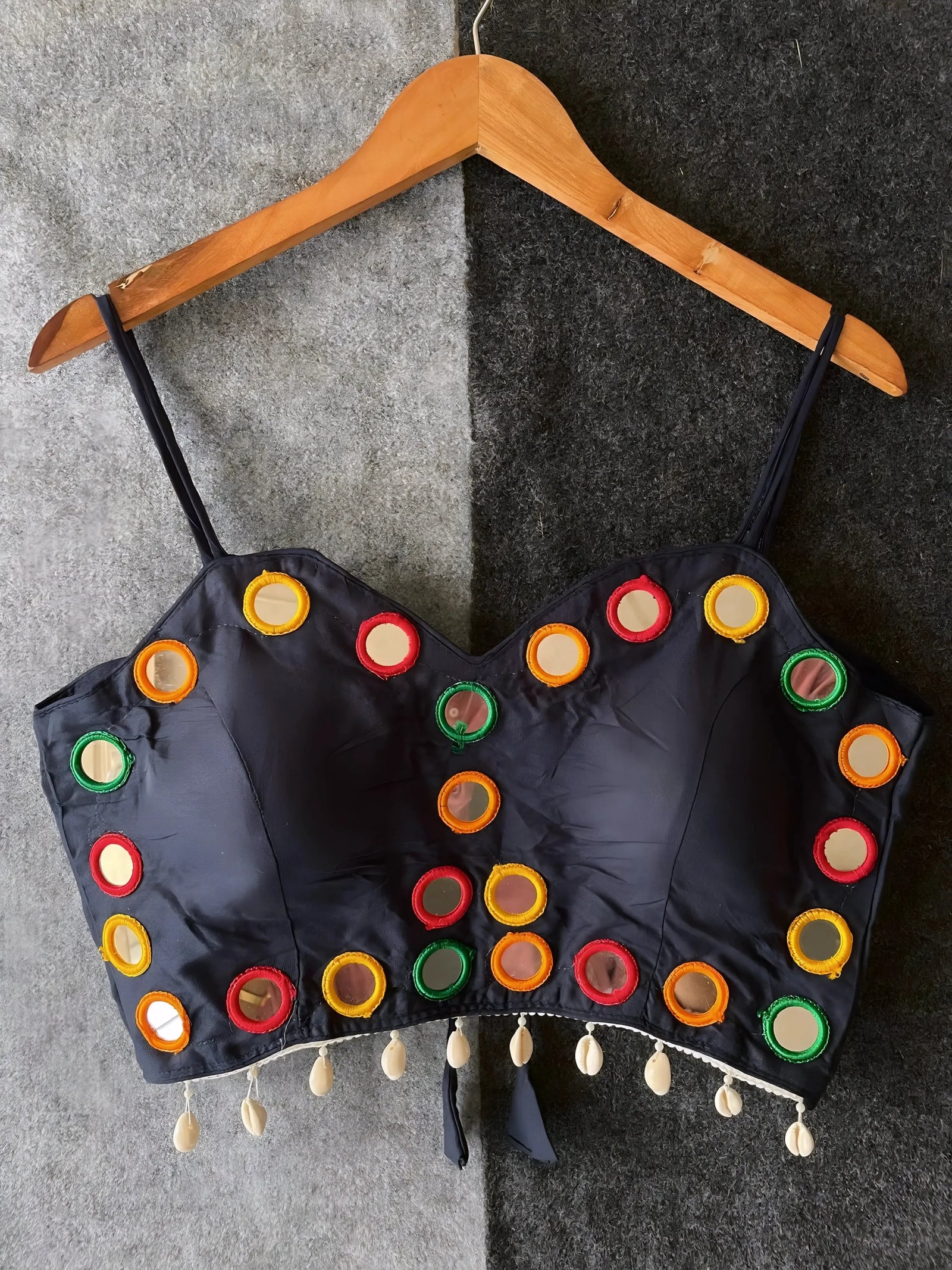 Navy corset-style blouse decorated with vibrant round mirrors and multicolored embroidered borders, featuring thin shoulder straps and shell tassel drops along the hem. Displayed on a wooden hanger against a two-tone grey textured background, highlighting its festive and contemporary ethnic design.