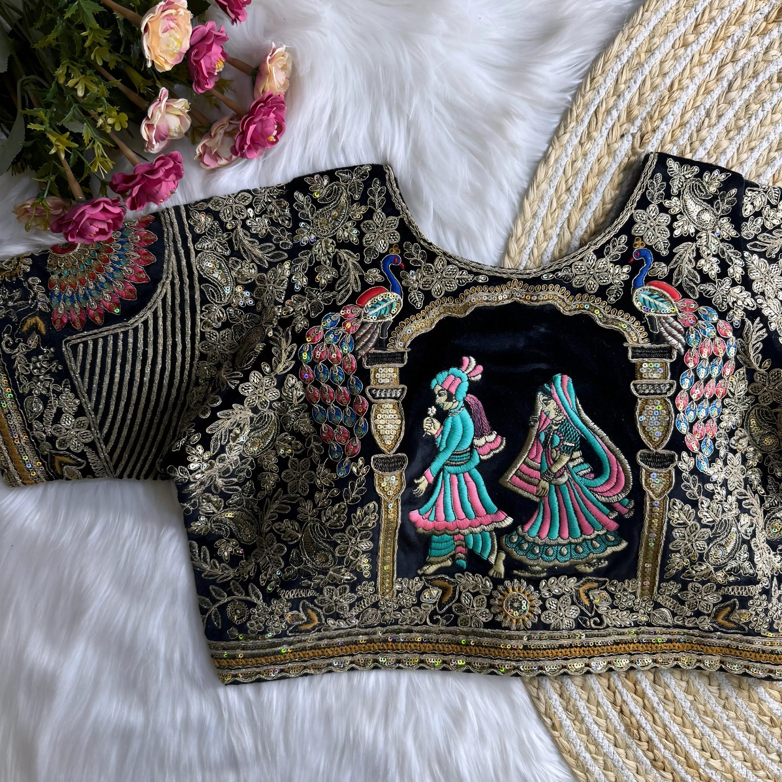 Traditional navy blue silk blouse featuring intricate gold zardosi embroidery with floral and leaf motifs, sequin embellishments, geometric panel designs, and a detailed multicolor peacock feather motif on the sleeve, displayed flat on a white fur surface with a braided mat and artificial pink flowers in the corner.