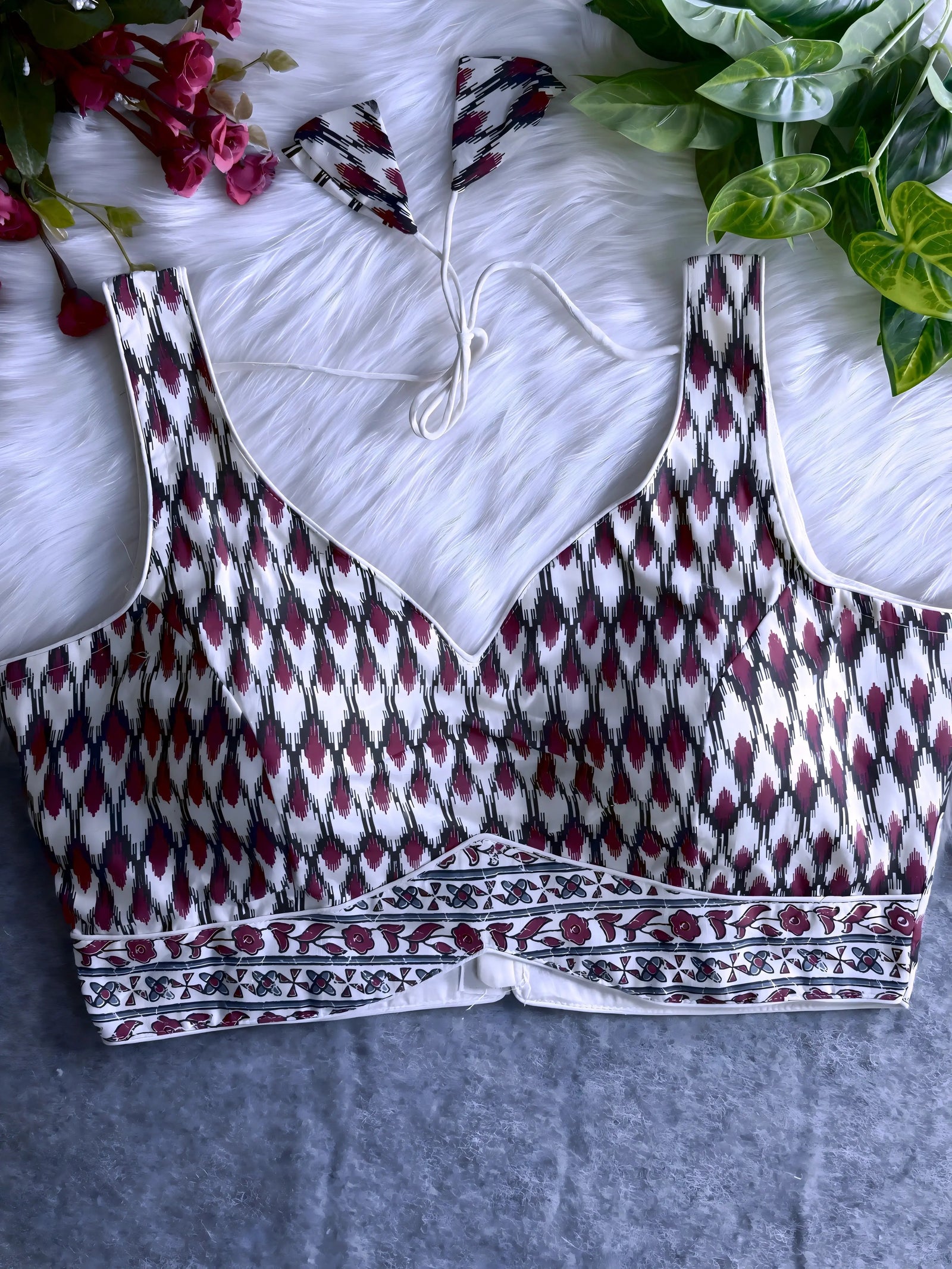 Off-white silk blouse featuring a maroon and black Ikat-inspired geometric print, wide shoulder straps, contoured cups, and a decorative floral motif border at the hem. The design includes a V-neckline and matching patterned tasseled tie-back, displayed flat on a white fur background with artificial pink flowers and green foliage at the corners.
