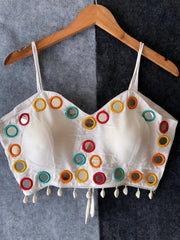 Off-white corset-style blouse adorned with vibrant round mirror work and multicolored embroidered borders, featuring thin shoulder straps and shell tassel drops along the hem. Displayed on a wooden hanger against a two-tone grey textured background, showcasing a festive and contemporary ethnic design.