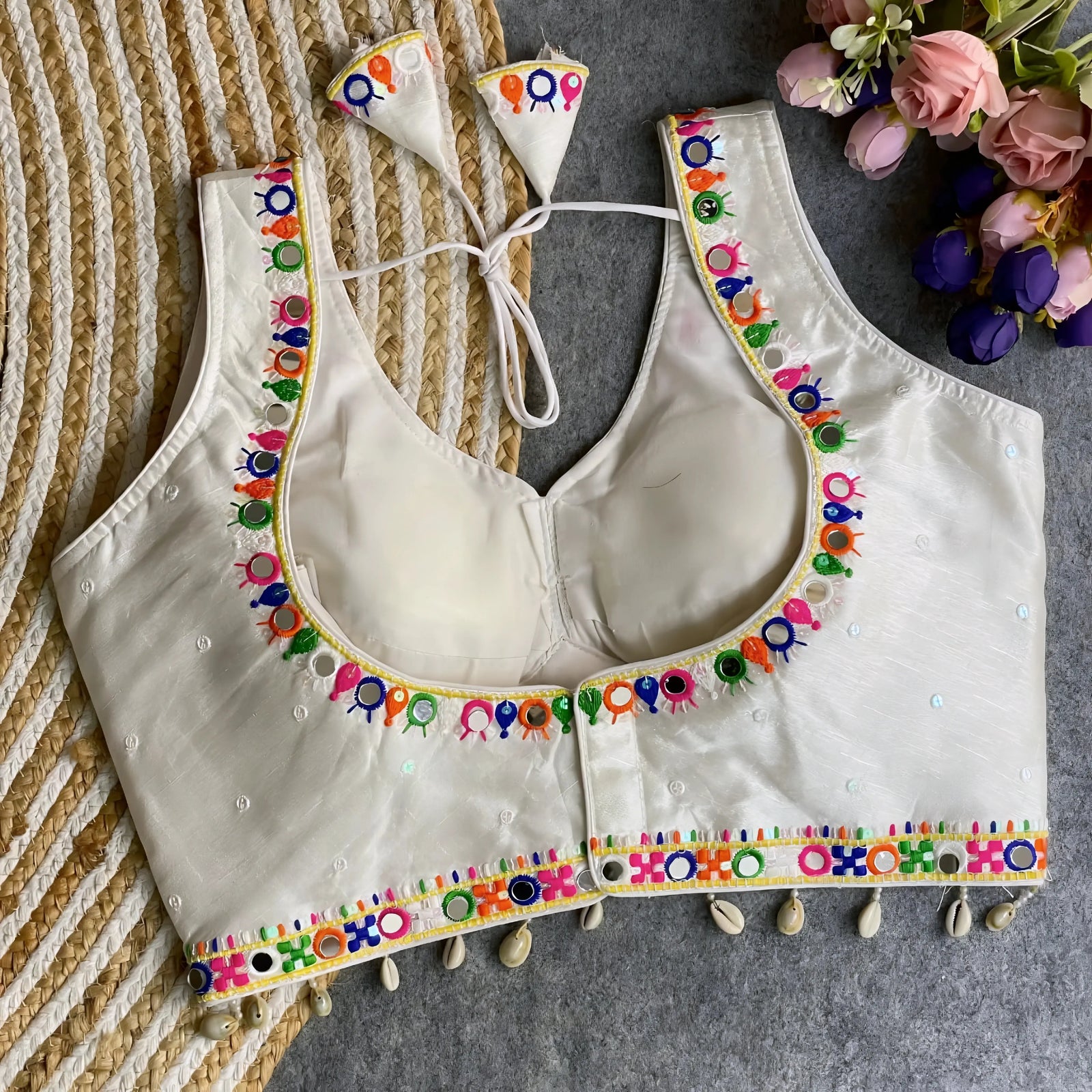 Off-white blouse featuring vibrant mirror work and traditional Kutch multicolor embroidery along the neckline and hem, with delicate sequin accents and shell tassels. The blouse includes a stylish back tie-up with matching embroidered tassels and is displayed flat on a textured surface with a floral bouquet, highlighting its festive ethnic elegance.