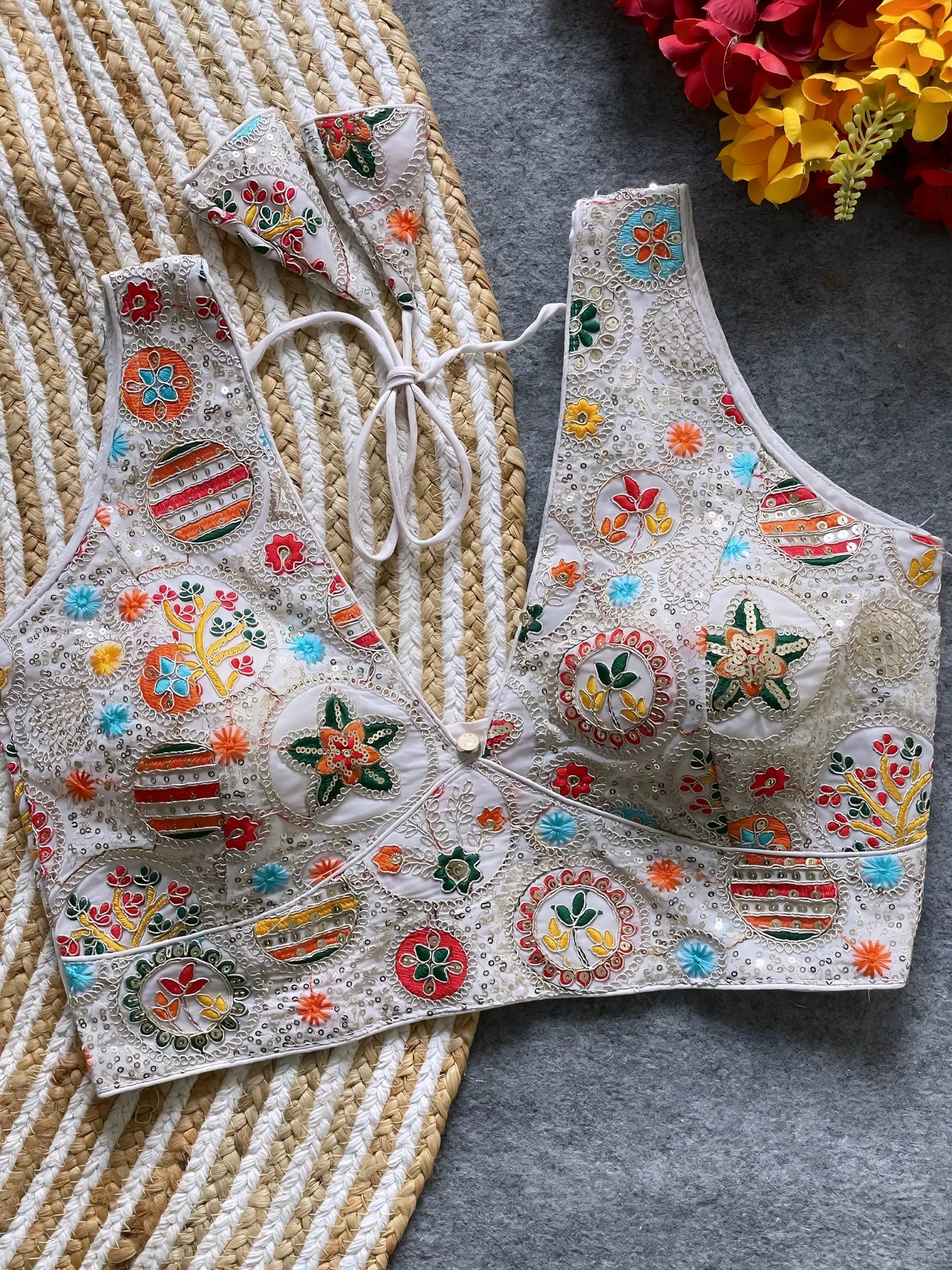 Traditional off-white blouse featuring vibrant multicolor thread embroidery and mirror work in circular floral and geometric patterns, with a deep V-neck and tie-back detail, displayed flat on a textured mat with artificial red and yellow flowers in the corner.
Related
Ethnic-saree-with intricate embroidery
Colorful saree fabric close-up
Traditional Madhubani art design
Vibrant handcrafted saree pattern.