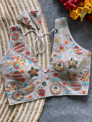 Traditional off-white blouse featuring vibrant multicolor thread embroidery and mirror work in circular floral and geometric patterns, with a deep V-neck and tie-back detail, displayed flat on a textured mat with artificial red and yellow flowers in the corner.
Related
Ethnic-saree-with intricate embroidery
Colorful saree fabric close-up
Traditional Madhubani art design
Vibrant handcrafted saree pattern.