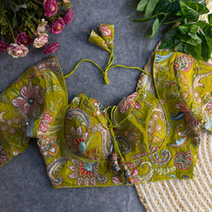Olive green padded blouse with intricate multicolor floral embroidery and paisley patterns, featuring a tie-back detail, displayed on a grey surface with artificial pink flowers and green leaves in the background.