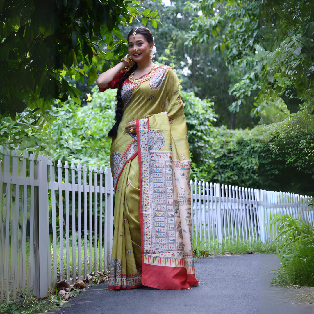 A woman models a striking Olive Green Soft Tussar Silk Saree with a red accent border. The pallu features a wide, elaborate panel with intricate White Madhubani-style folk art prints and a solid red hem. She wears a red blouse and a heavy gold choker necklace, posing outdoors.