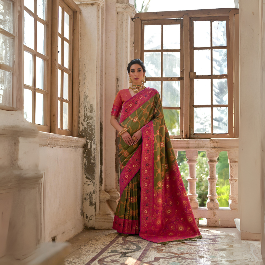 A woman models a striking Mehandi Green Banarasi Silk Saree with gold Zari weaving. The saree has a wide, ornate Fuchsia Pink border and pallu featuring dense traditional geometric Zari weaving. She wears a coral pink blouse and heavy gold Kundan jewelry.