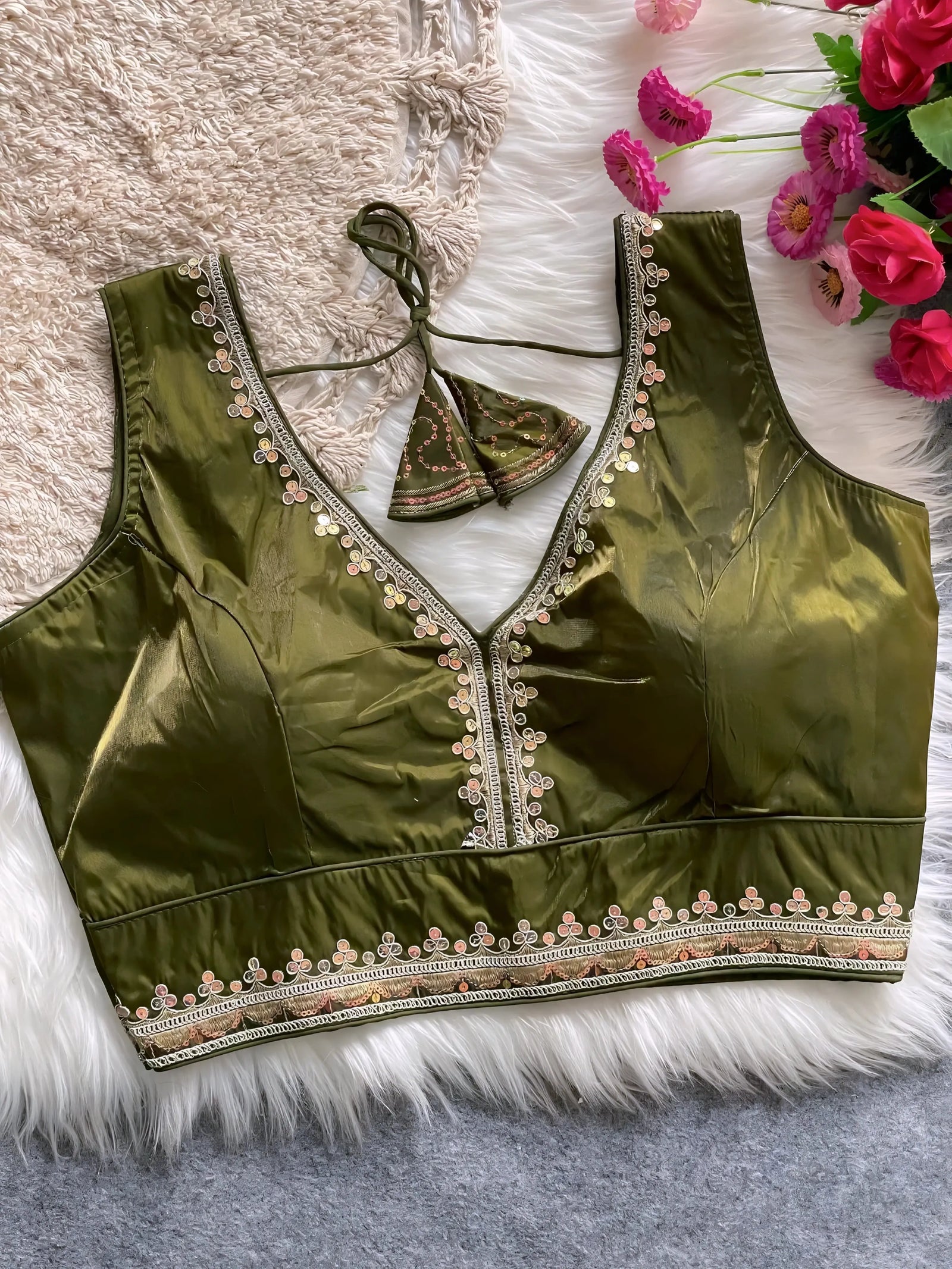 Traditional olive green silk blouse featuring intricate pastel embroidery along the neckline and hem, with a decorative tassel tie at the back, displayed flat on a soft fur and textured mat background with artificial pink flowers in the corner.