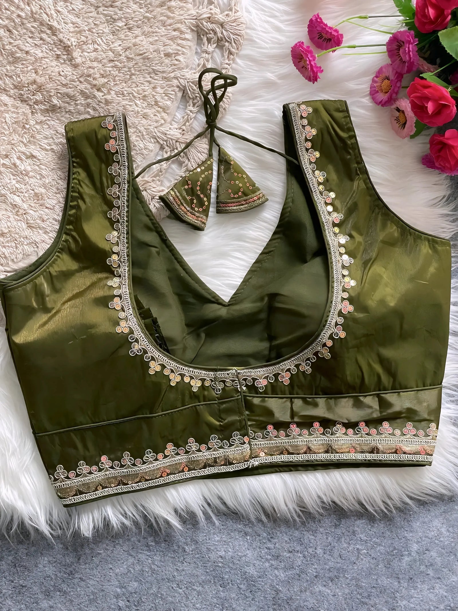 Traditional olive green silk blouse featuring intricate pastel embroidery along the neckline and hem, with a decorative tassel tie at the back, displayed flat on a soft fur and textured mat background with artificial pink flowers in the corner.