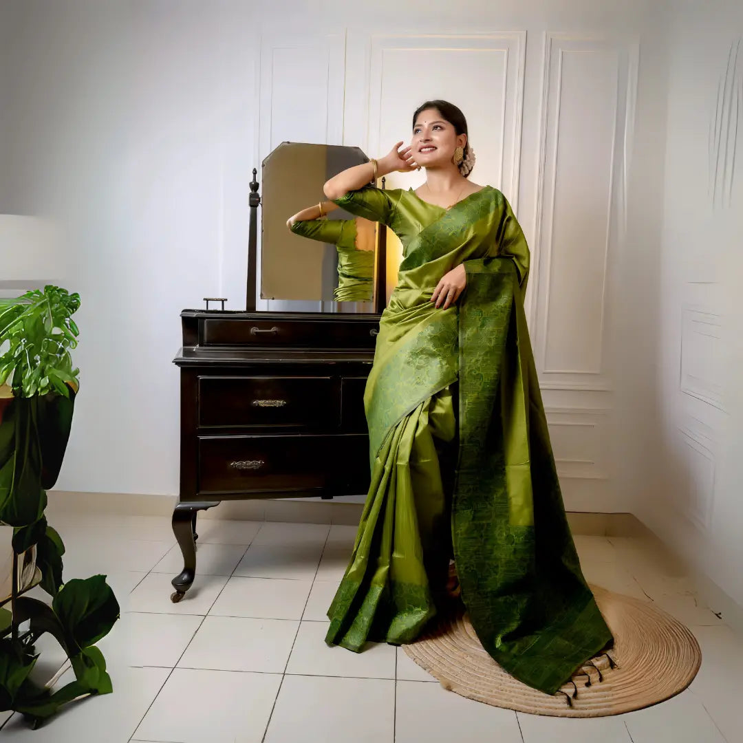 A woman models a luxurious Olive Green Raw Silk Saree. The saree features a wide border and pallu with dense, intricate tone-on-tone Kalamkari-style weaving in a matching green Zari. She wears a long-sleeve green blouse and subtle gold jewelry, with white flowers adorning her hair, posing indoors next to a dark wooden vanity.