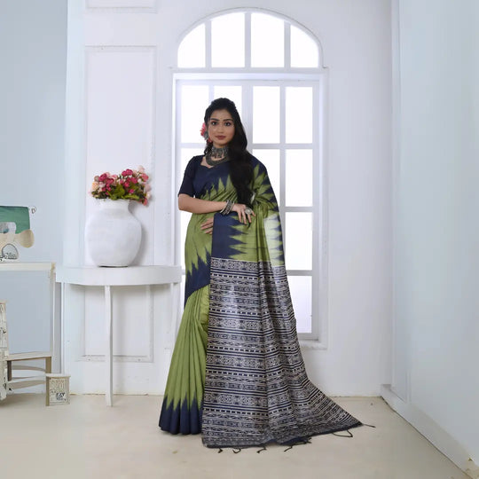 A woman models a striking Pista Green Soft Tussar Silk Saree. The saree features a bold black/navy zigzag or V-shaped pattern along the border and transitions on the body. The pallu has a wide panel of dense tribal or Ikat-style geometric print in light grey and white on a dark background. She wears a black/navy blouse and chunky oxidized silver jewelry, posing indoors by a large arched window.