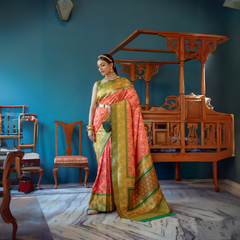 A woman models a striking Peach/Coral Banarasi Silk Saree with all-over Zari weaving. The wide pallu and border contrast dramatically in Olive Green with large geometric Zari weaving. She wears a green blouse, traditional gold jewelry, and a metallic waist belt, posing in an ornate room.
