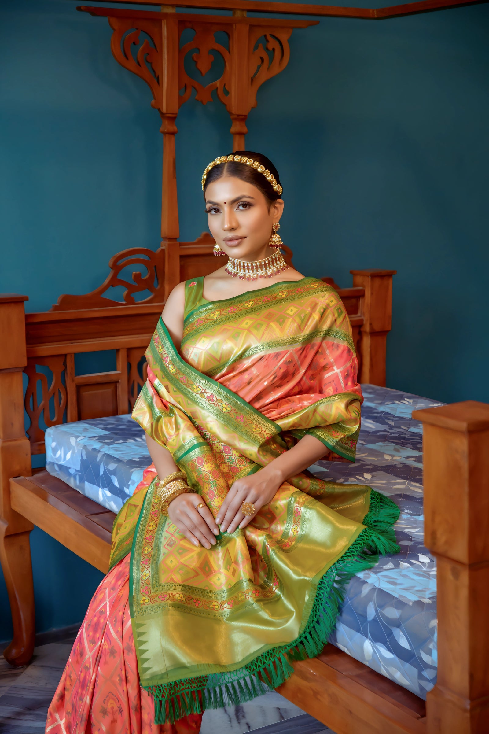 A woman models a striking Peach/Coral Banarasi Silk Saree with all-over Zari weaving. The wide pallu and border contrast dramatically in Olive Green with large geometric Zari weaving. She wears a green blouse, traditional gold jewelry, and a metallic waist belt, posing in an ornate room.