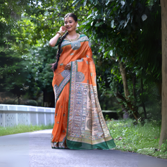 A woman models a vibrant Coral Orange Soft Tussar Silk Saree. The saree has a deep green border and an elaborate pallu featuring intricate White Madhubani-style folk art prints with blue and green figurative motifs. She wears a green embellished blouse and a gold Kundan choker necklace.