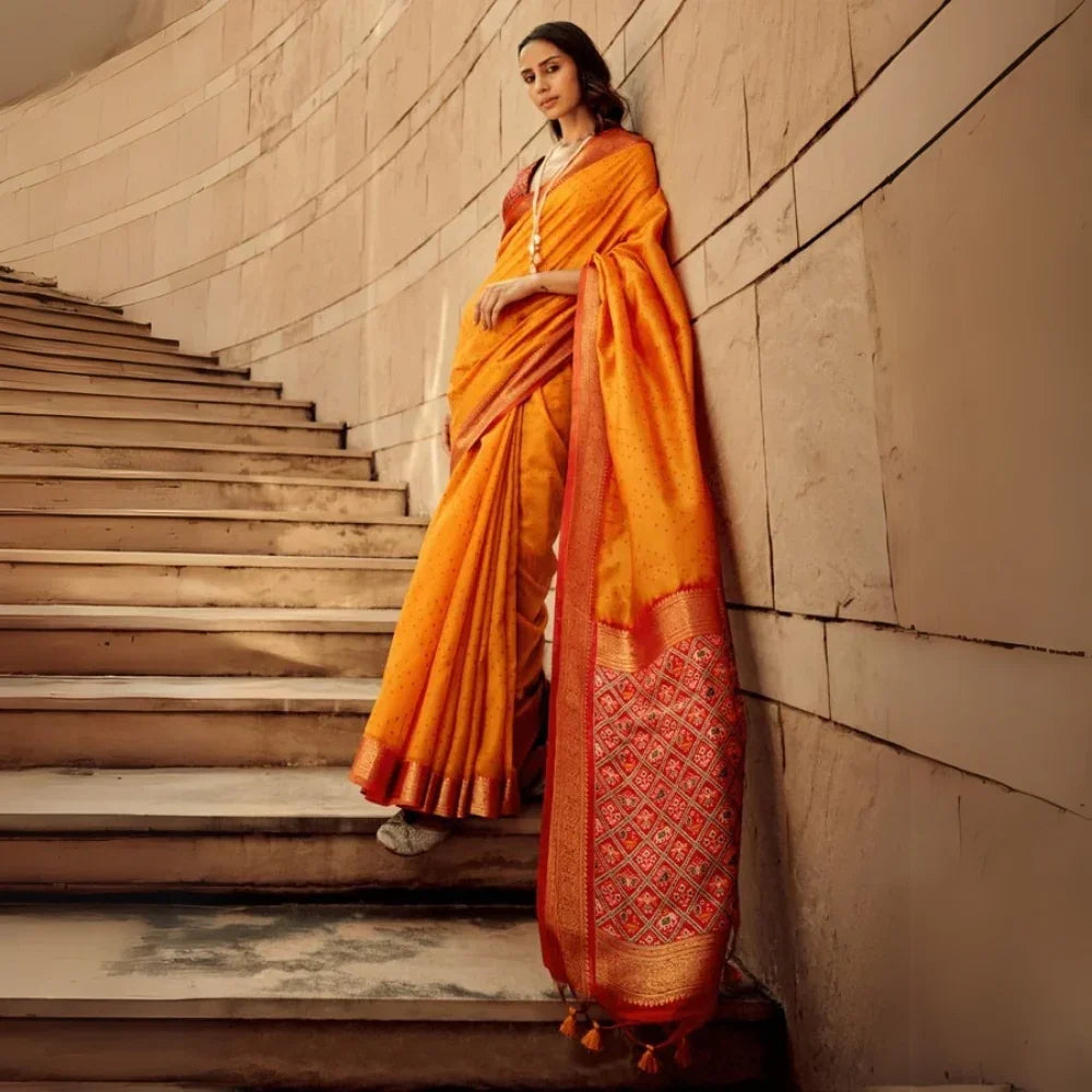 Woman wearing elegant orange red saree with intricate zari work and rich pallu design, standing on stone stairs in ethnic fashion look