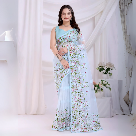 A woman models an ethereal Light Blue Soft Net Saree. The sheer fabric is densely covered in delicate all-over floral embroidery featuring white, pink, red, and green threadwork. She wears a matching sleeveless light blue blouse and accessorizes with sophisticated silver/diamond jewelry, posing indoors against sheer white curtains.