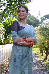 A woman models an elegant Sky Blue Soft Muga Cotton Saree. The pallu features large, artistic Resham floral motifs in shades of blue and indigo. She wears a contrasting navy blouse and is accessorized with a pearl choker necklace and vibrant orange bangles, posing outdoors by a white fence.