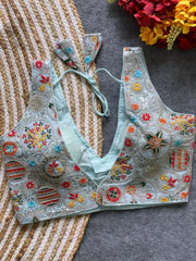 Pastel blue blouse featuring vibrant multicolor thread embroidery and mirror work in floral and geometric patterns, with sequin accents and a deep V-neck, displayed flat on a textured mat with artificial red and yellow flowers in the corner.