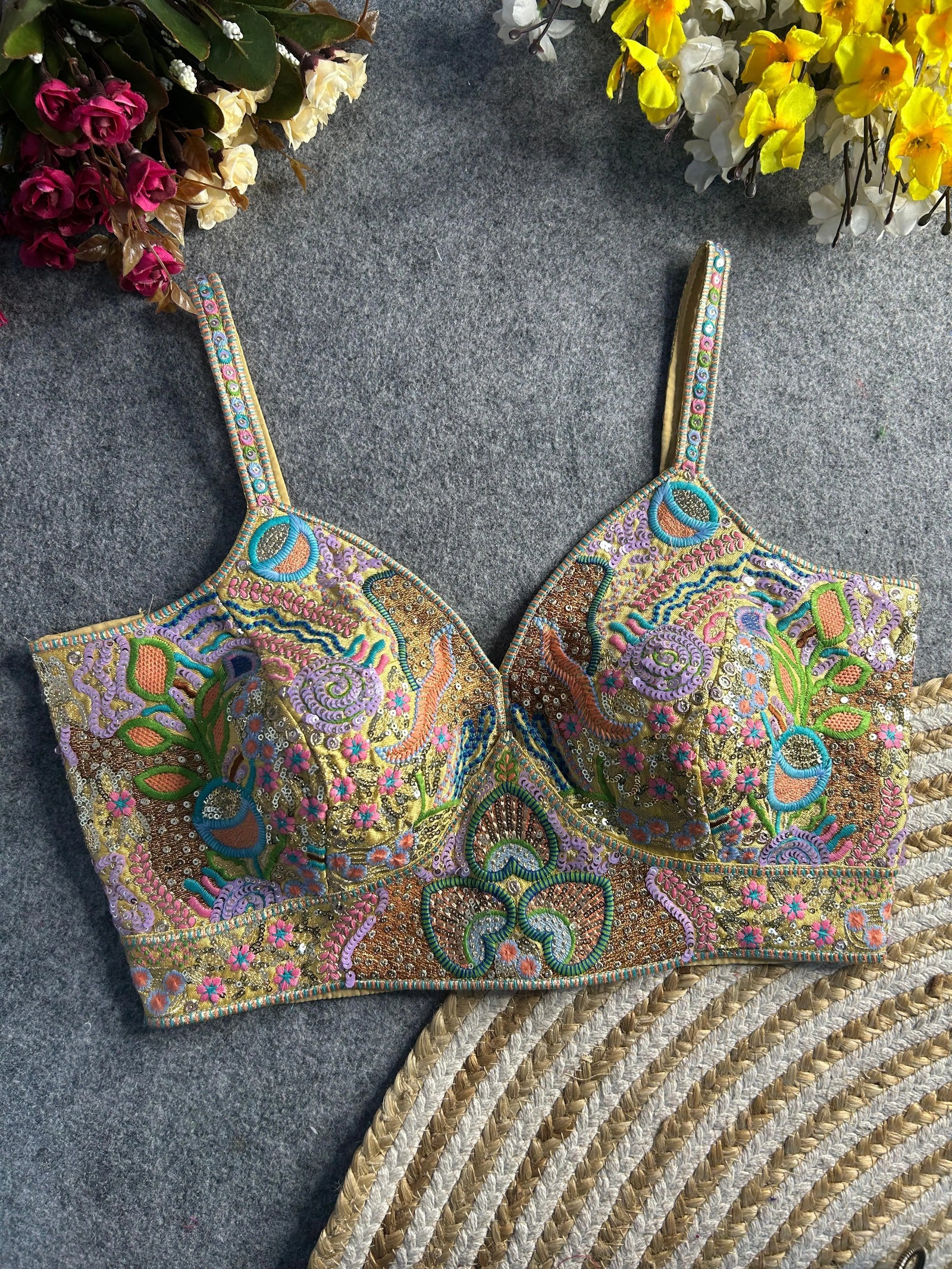Pastel multicolor bustier-style blouse featuring intricate thread embroidery and sequin embellishments in floral, paisley, and geometric motifs, with structured cups and adjustable straps, displayed flat on a textured grey surface surrounded by artificial pink, yellow, and white flowers in the corners.
