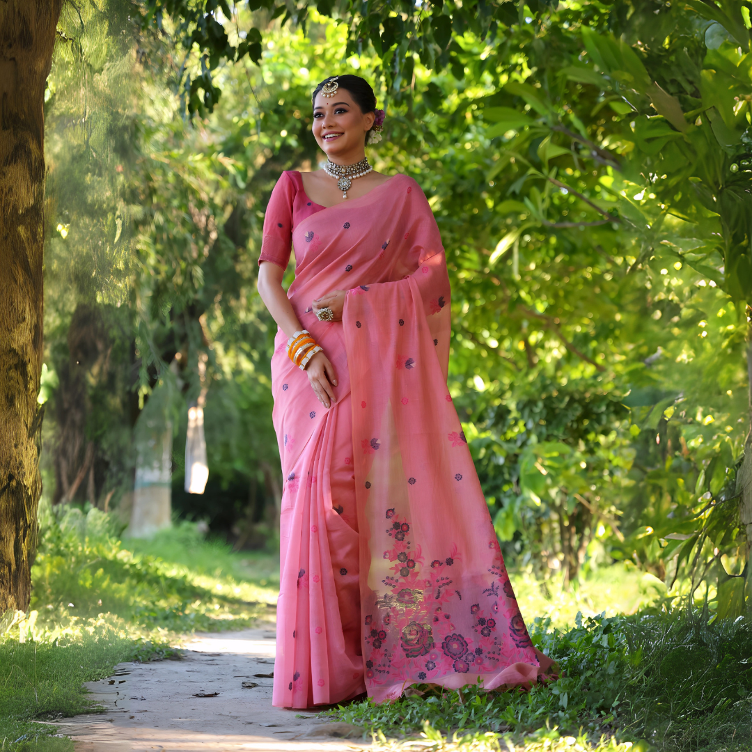 A woman models an elegant light Pink Soft Muga Cotton Saree. The body has scattered floral Resham weaving, and the pallu features an elaborate panel of darker floral weaving/print in purple and blue tones. She wears a matching pink blouse, white stone/Kundan jewelry, and vibrant orange and yellow bangles, posing outdoors.