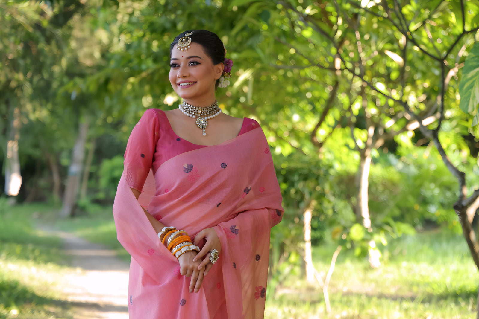 A woman models an elegant light Pink Soft Muga Cotton Saree. The body has scattered floral Resham weaving, and the pallu features an elaborate panel of darker floral weaving/print in purple and blue tones. She wears a matching pink blouse, white stone/Kundan jewelry, and vibrant orange and yellow bangles, posing outdoors.