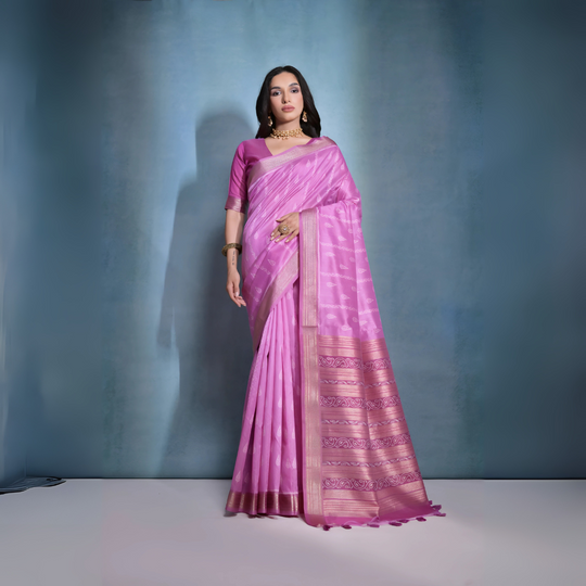 A woman models a stunning Deep Pink Soft Raw Silk Saree. The saree features subtle tone-on-tone Ikat weaving on the body, framed by a wide, luxurious border of dark magenta and intricate gold Zari weaving. She wears a matching pink blouse and gold Kundan jewelry, posing indoors against a textured blue backdrop.