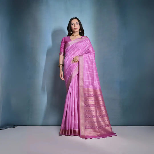 A woman models a stunning Deep Pink Soft Raw Silk Saree. The saree features subtle tone-on-tone Ikat weaving on the body, framed by a wide, luxurious border of dark magenta and intricate gold Zari weaving. She wears a matching pink blouse and gold Kundan jewelry, posing indoors against a textured blue backdrop.