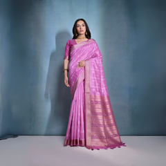 A woman models a stunning Deep Pink Soft Raw Silk Saree. The saree features subtle tone-on-tone Ikat weaving on the body, framed by a wide, luxurious border of dark magenta and intricate gold Zari weaving. She wears a matching pink blouse and gold Kundan jewelry, posing indoors against a textured blue backdrop.