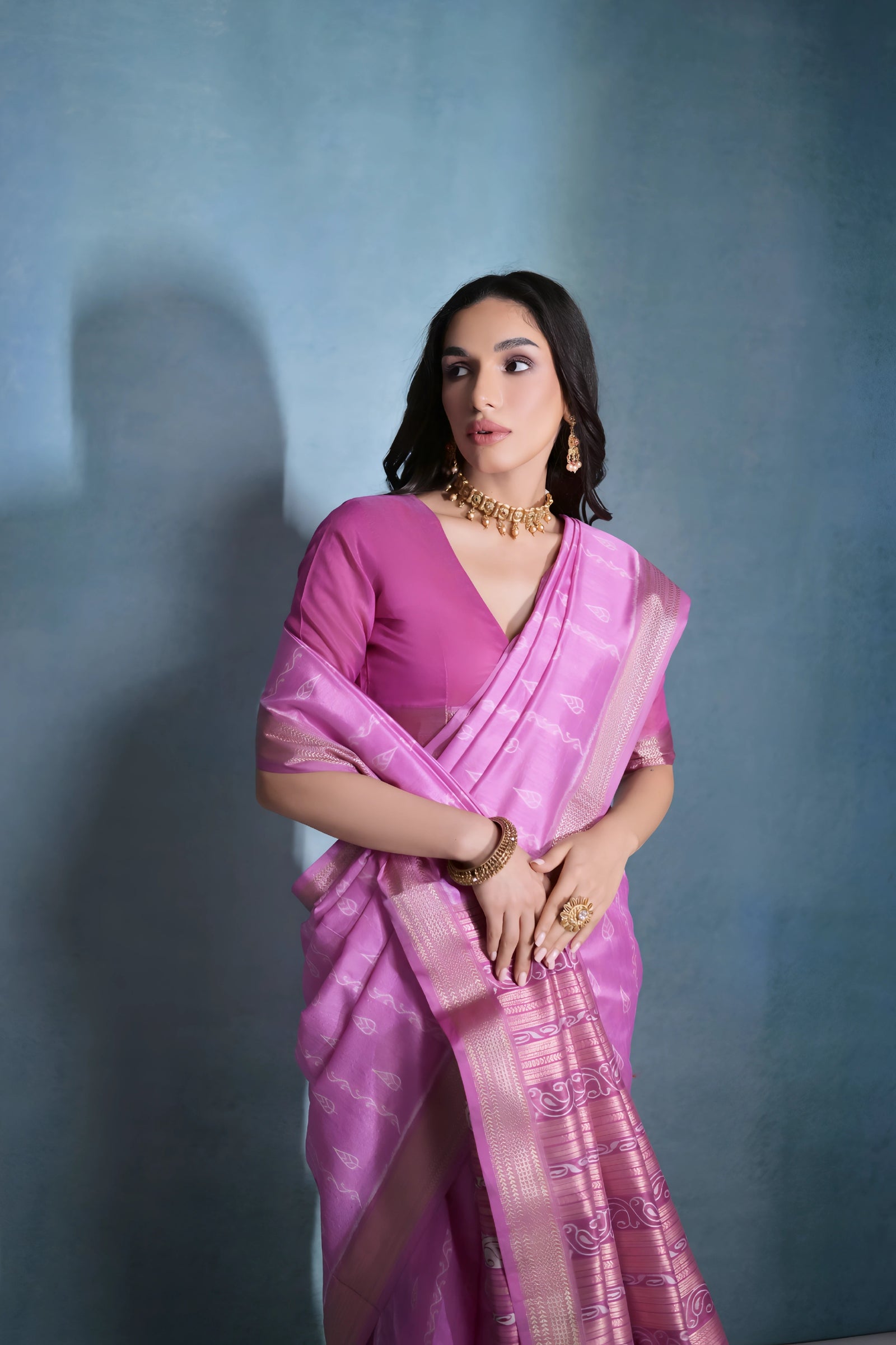 A woman models a stunning Deep Pink Soft Raw Silk Saree. The saree features subtle tone-on-tone Ikat weaving on the body, framed by a wide, luxurious border of dark magenta and intricate gold Zari weaving. She wears a matching pink blouse and gold Kundan jewelry, posing indoors against a textured blue backdrop.