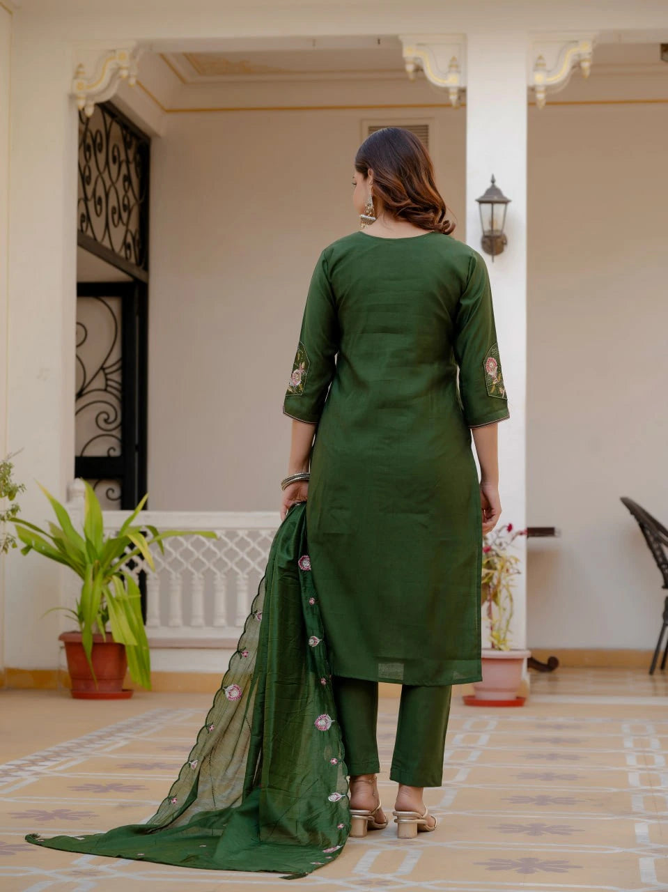 Woman in a green traditional outfit standing in a decorated room with plants and a lamp.