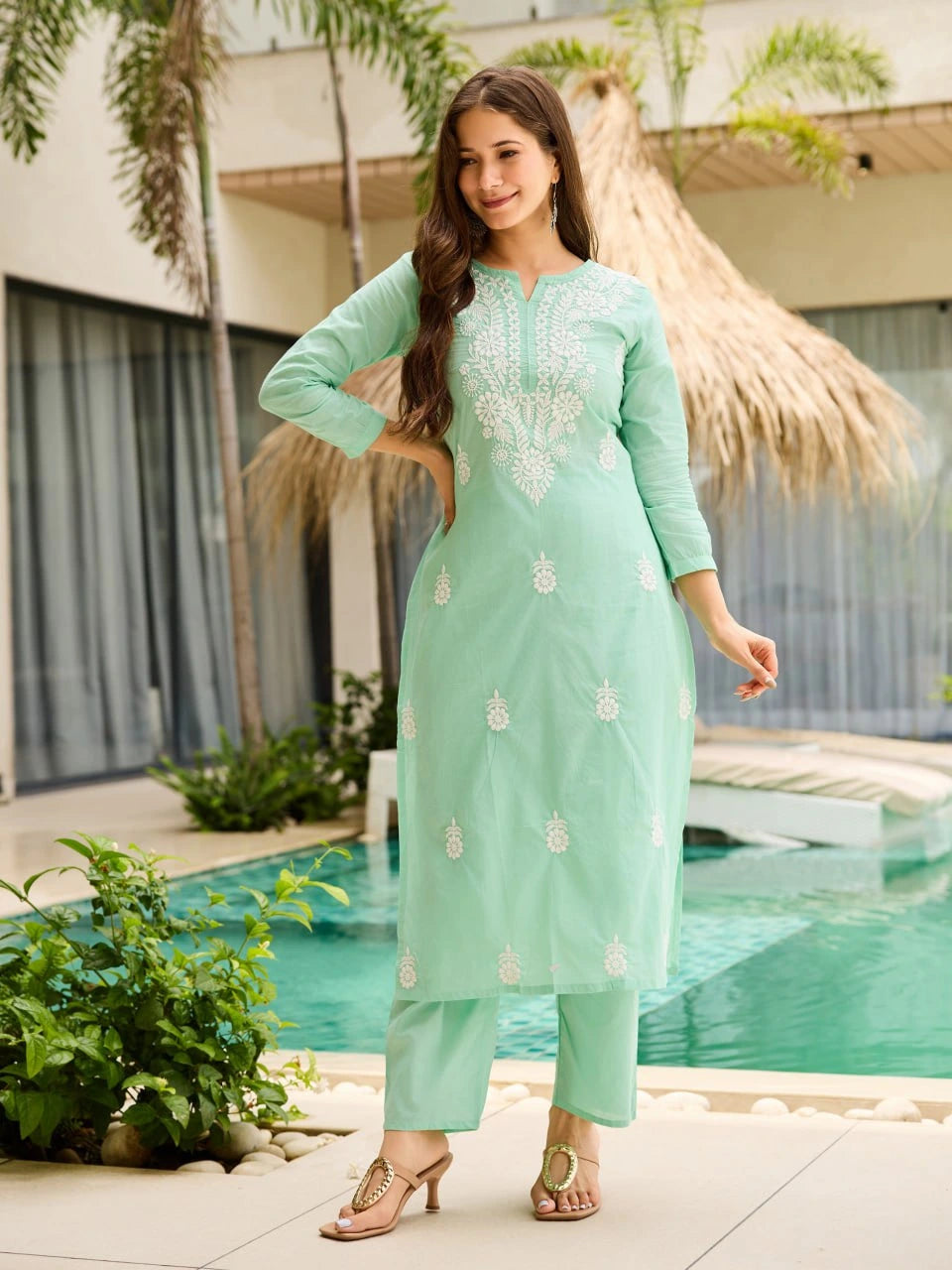 Woman in a light green traditional outfit standing by a pool with a modern building and palm trees in the background.