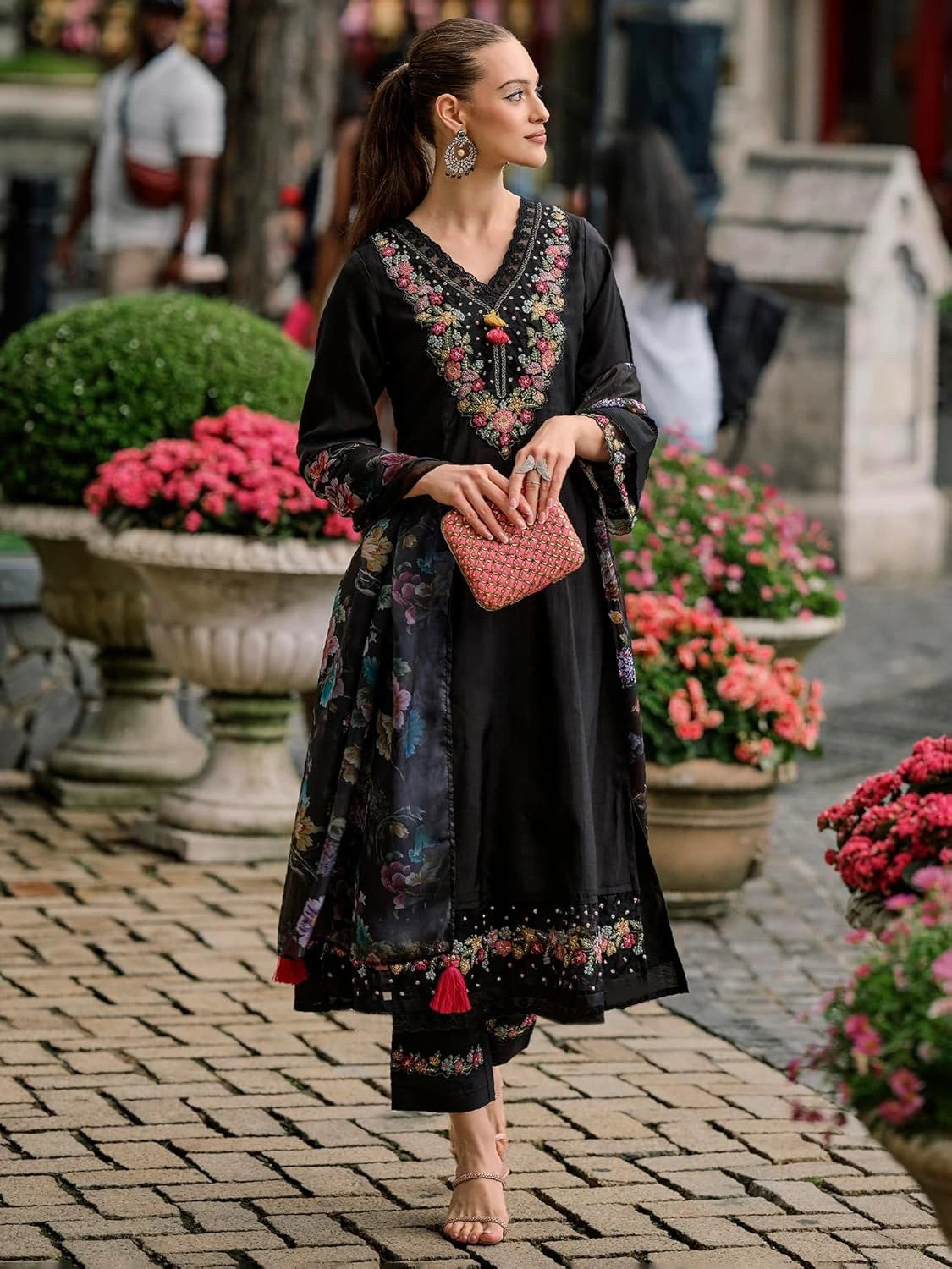 Woman in traditional black and floral outfit with pink clutch standing in a garden.