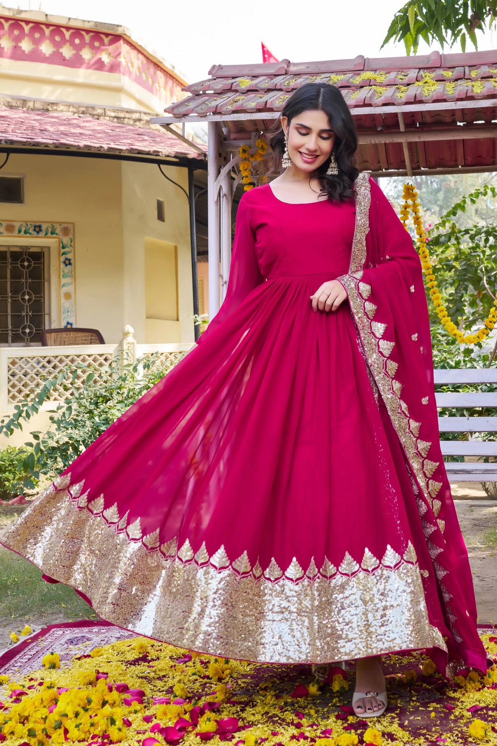 Woman in a pink traditional outfit with gold details standing in front of a house with greenery.