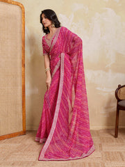 Woman wearing a pink saree with a white border in a room with a textured wall and wooden chair.