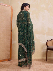 Woman in a green embroidered traditional outfit standing in a room with a textured wall and wooden chair.