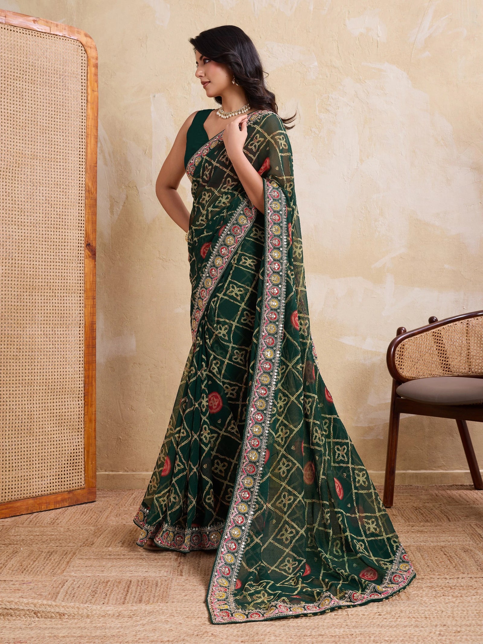 Woman wearing a green saree with a patterned border in a room with a textured wall and wooden chair.
