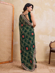Woman wearing a green saree with a pattern, standing in a room with a textured wall and wooden chair.