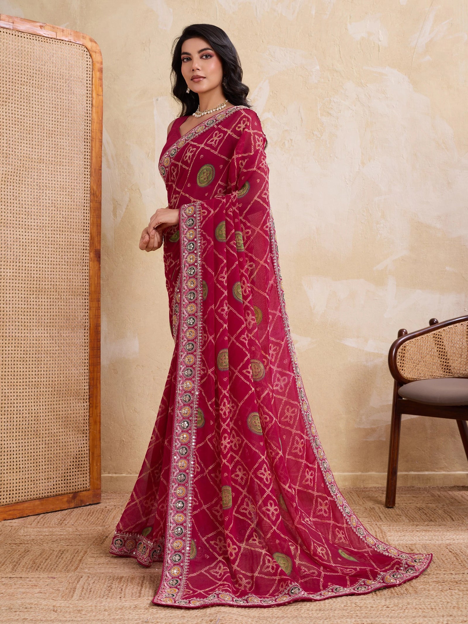Woman wearing a red saree with intricate patterns in a room with a textured wall and chair.