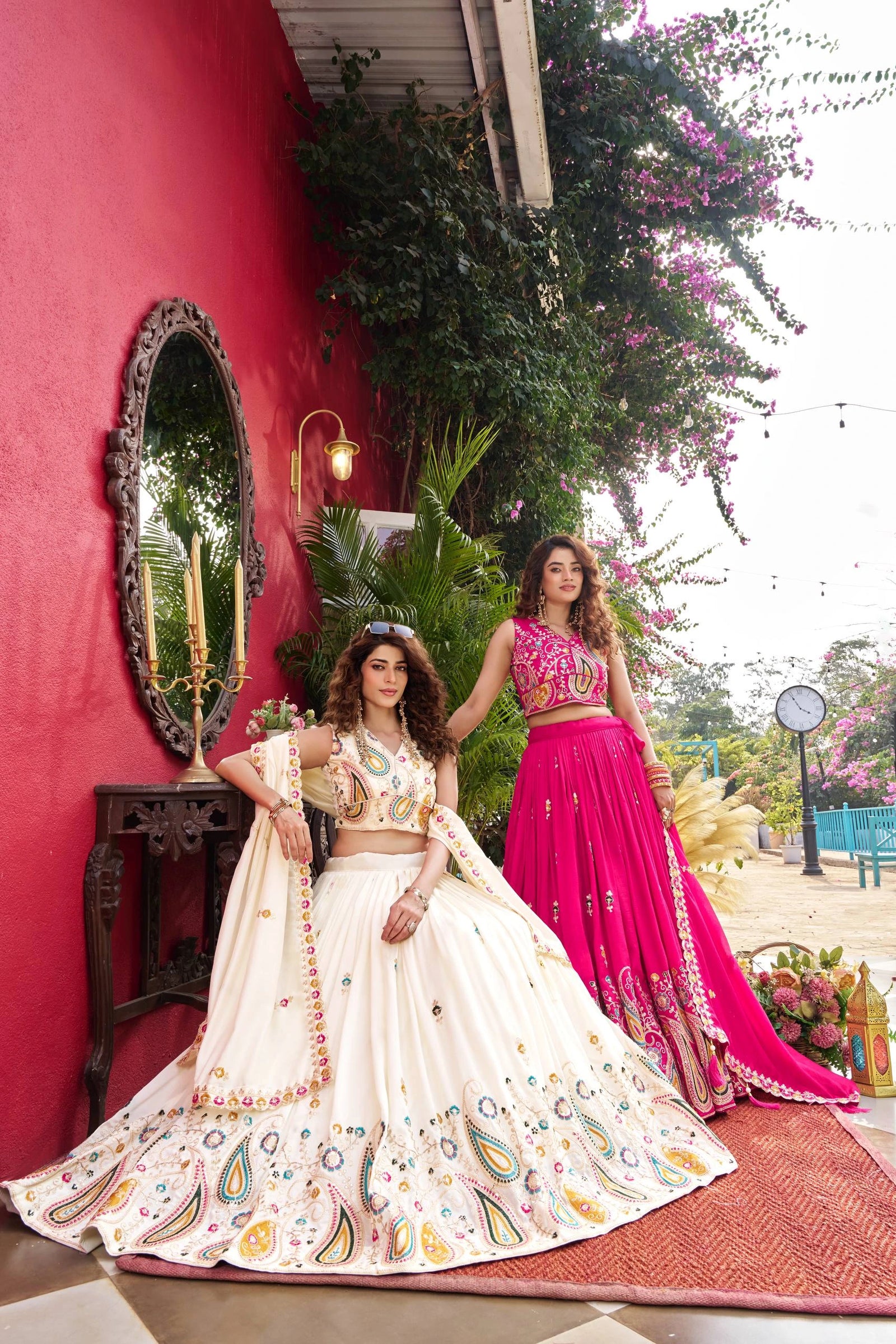 Two women in traditional outfits standing in front of a red wall with decorative elements.