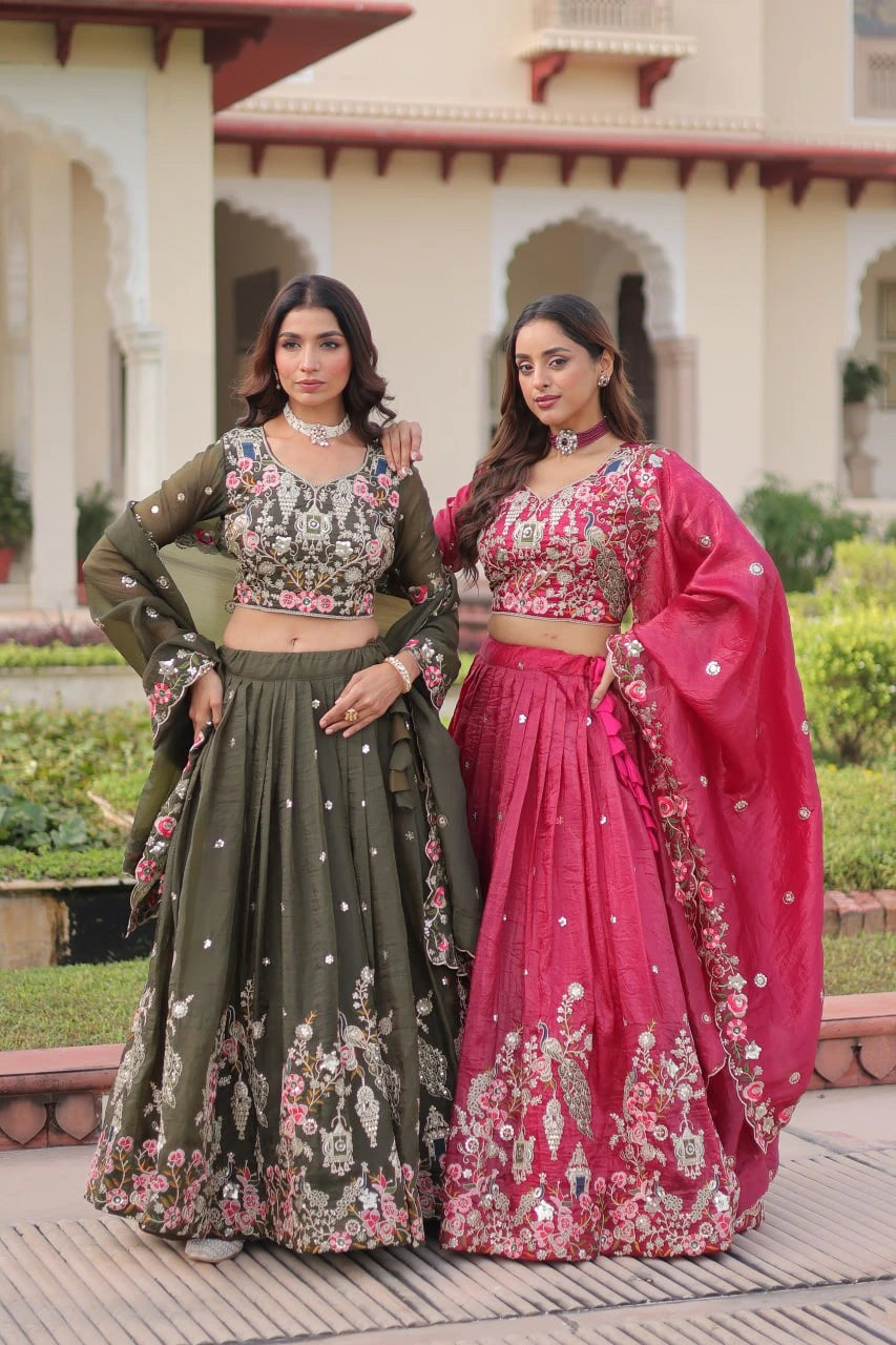 Two women in traditional outfits standing outdoors with a building and greenery in the background.