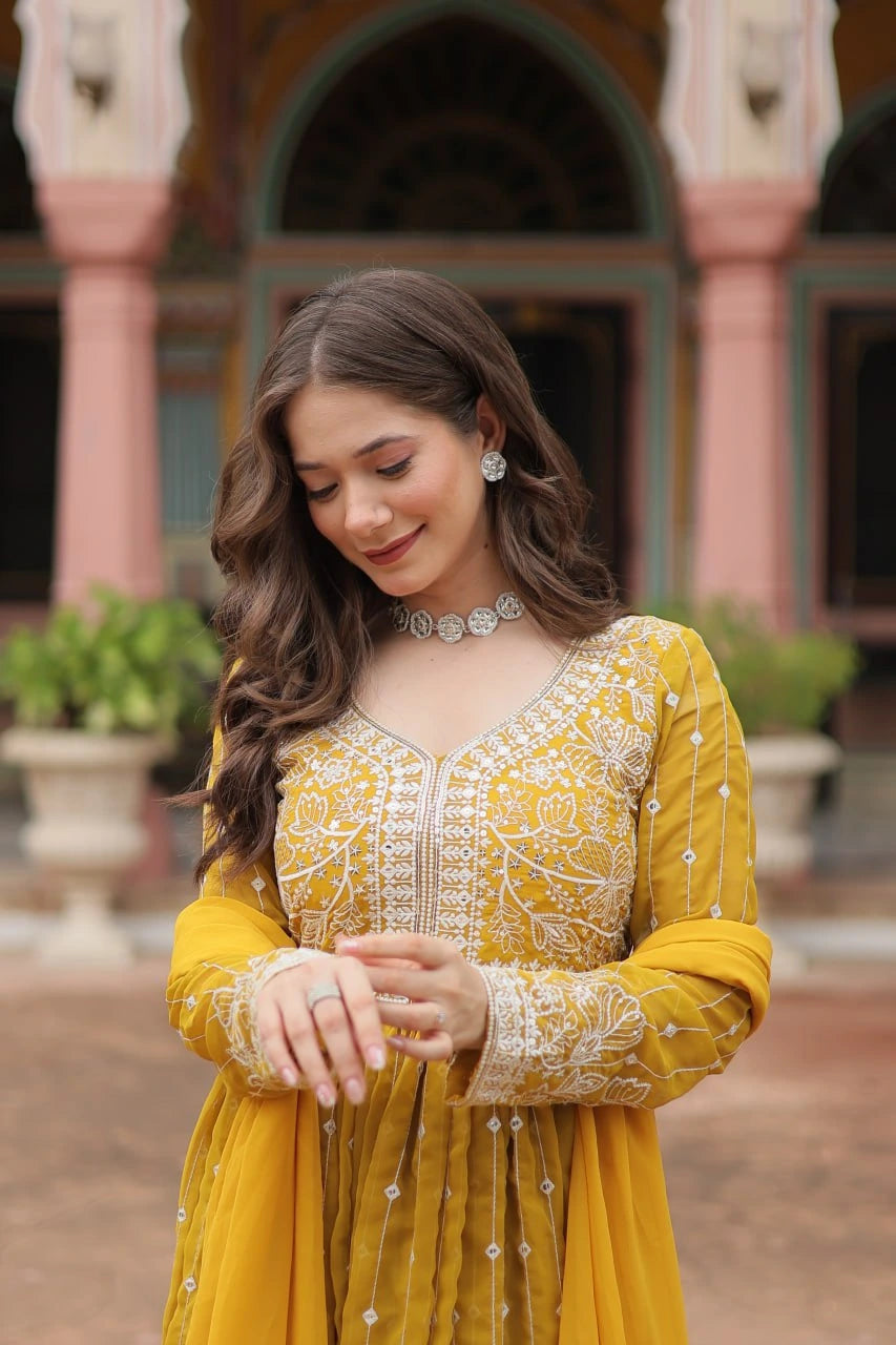 Woman in a yellow traditional outfit with intricate patterns standing in front of an architectural background.