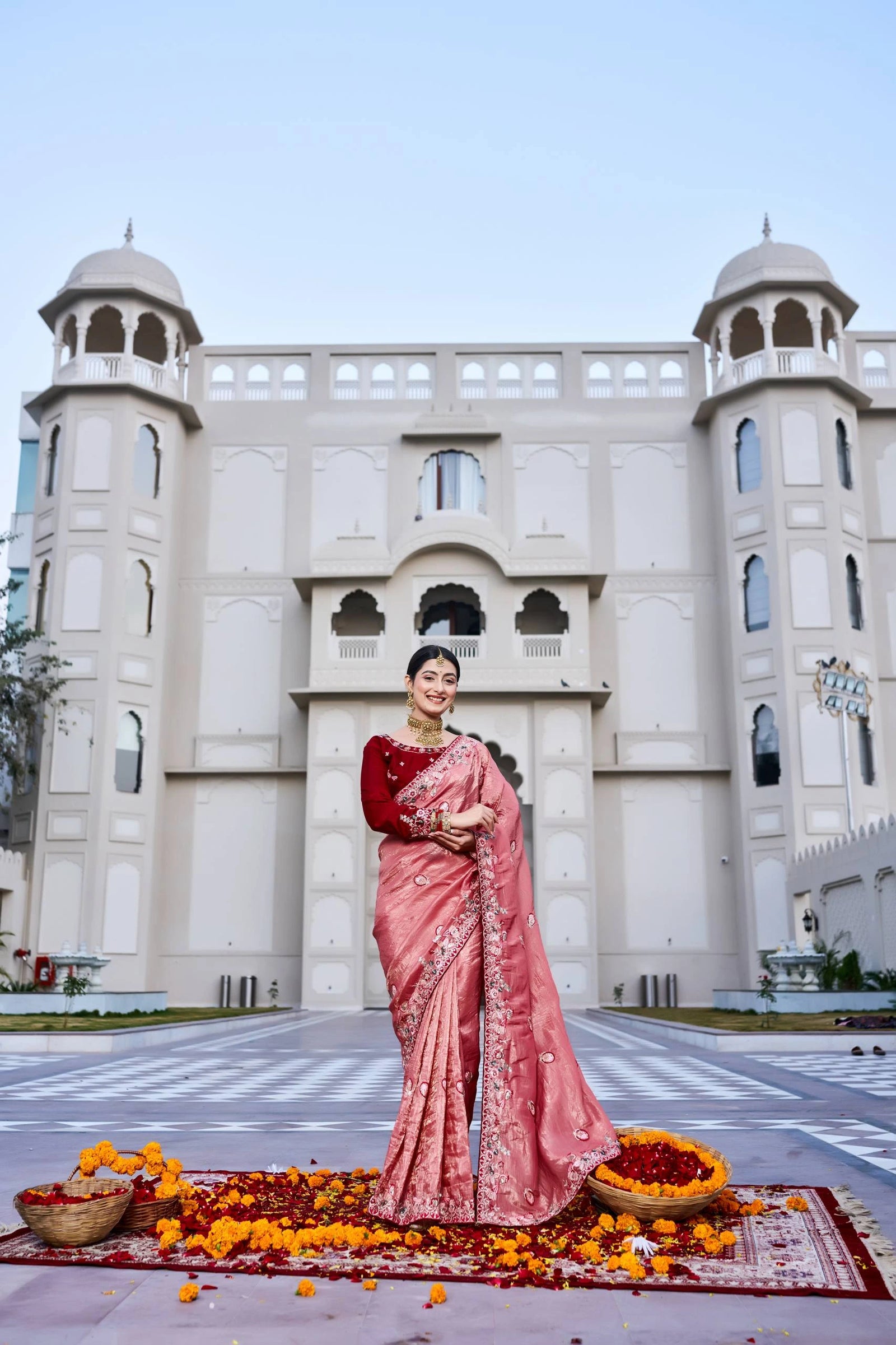 Woman in a pink saree standing in front of a large building with a blue sky.