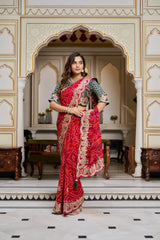 Woman in a red saree with intricate patterns standing in an ornate room.
