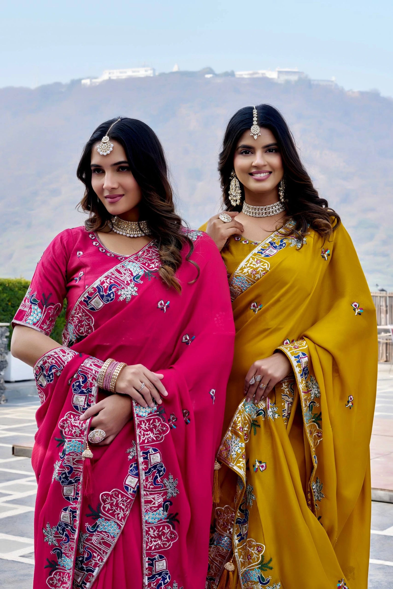 Two women in traditional sarees standing outdoors with a mountainous background.