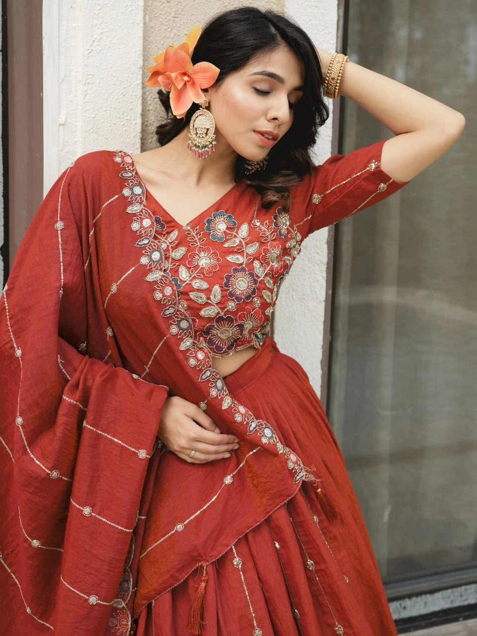 Woman in a red traditional outfit with floral embroidery, standing against a neutral wall.