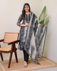Woman holding a black and white patterned dupatta in a room with a chair and plant.