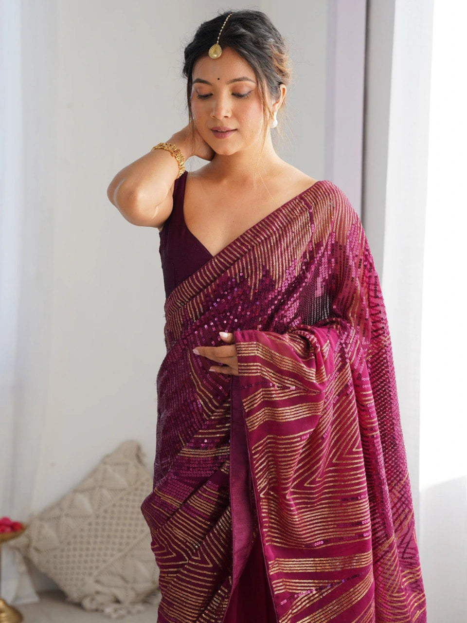 Woman wearing a maroon saree with gold patterns in a bright room.