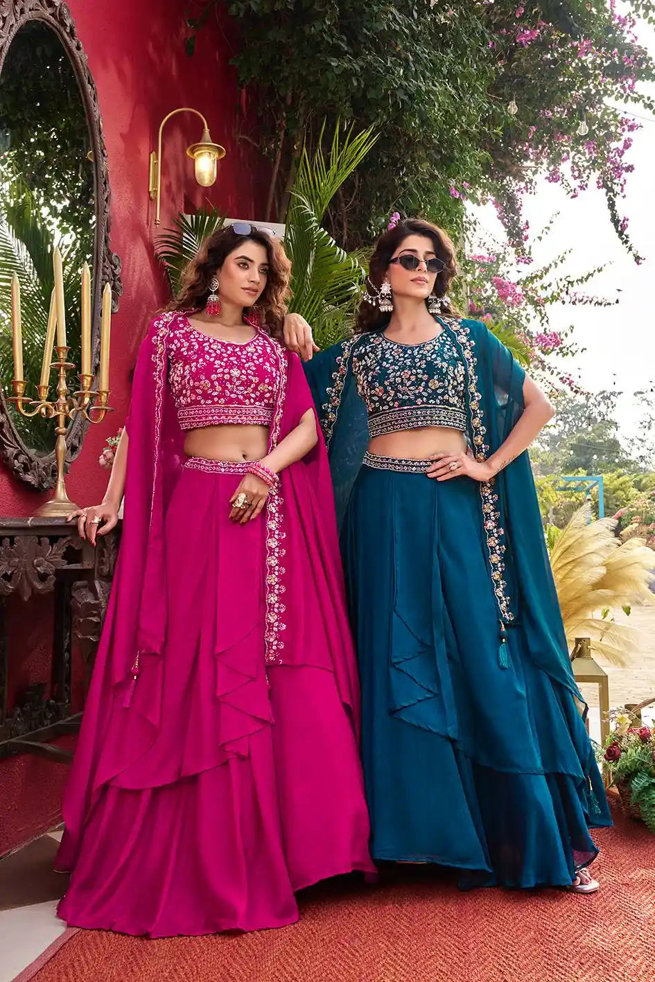 Two women in traditional pink and blue lehengas standing in a decorative outdoor setting.
